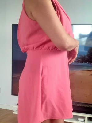 Rosa ärmlös jumpsuit med v-ringning - Säljer en snygg rosa jumpsuit med djup v-ringning och lös passform upptill. Den har resår i midjan och vida shorts. Perfekt för dig som vill ha en bekväm men trendig look. Materialet känns mjukt och följsamt, troligen polyester eller bomullsblandning.