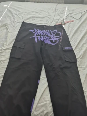 Svarta cargobyxor Minus Two graffiti - Säljer ett par svarta cargobyxor från Minus Two med lila graffiti-tryck på framsidan och fickan. Byxorna har stora sidofickor och bred passform, perfekta för en streetwear-look. Materialet känns slitstarkt och byxorna har bälteshällor.