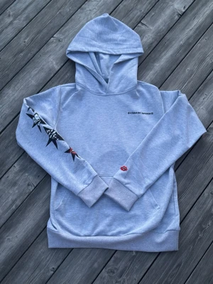 Ljusgrå chrome hearts hoodie - Ljusgrå hoodie från chrome hearts med coola tryck på ärmarna, bland annat stjärnor och döskallar på ena ärmen samt logga på den andra. Liten röd detalj vid mudden. Perfekt för dig som gillar streetstyle och unika detaljer.
