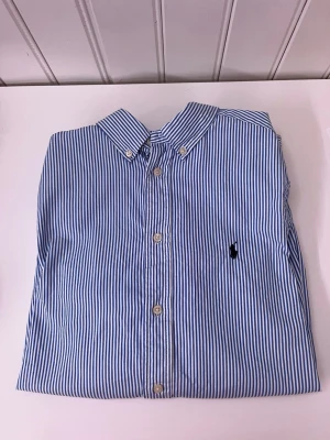 Polo Ralph Lauren skjorta - Riktigt snygg blårandig skjorta från Polo Ralph Lauren med vita knappar och mörkblått polo märke på bröstet.