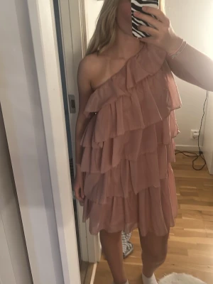 Rosa one shoulder volangklänning - Säljer en superfin rosa kortklänning med flera lager volanger och one shoulder-design. Klänningen har en luftig passform och är perfekt för sommarens alla tillfällen. Materialet känns lätt och flowy, och färgen är en mjuk rosa ton.