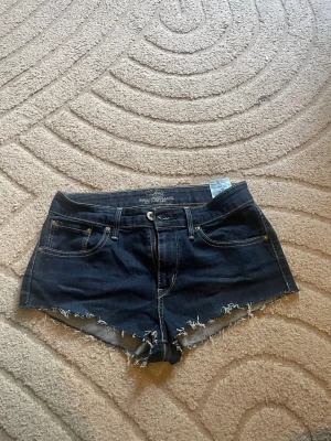 Mörkblå Levi's jeansshorts med rå kant - Snygga mörkblå jeansshorts från So med rå, fransig kant och klassisk femficksdesign. Shortsen har låg midja och kontrastsömmar för en cool look. Perfekta för varma dagar och enkel att matcha med olika toppar.