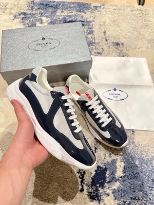 Prada sneakers i marinblå och vit - Snygga sneakers från Prada i marinblått och vitt med vita skosnören och röda detaljer på plösen. Ovandelen är i en mix av glansigt läder och mesh-tyg för en modern look. Sulan är vit och chunky, vilket ger en sportig vibe. Perfekta för dig som vill ha något exklusivt och trendigt.