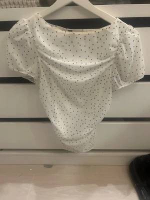 Vit prickig topp från H&M Divided - Supersöt vit topp från H&M Divided med små svarta prickar och puffiga korta ärmar. Toppen har en lätt rynkad effekt vid axlarna och är gjord i ett mjukt polyestertyg. Perfekt för en trendig och lekfull look!