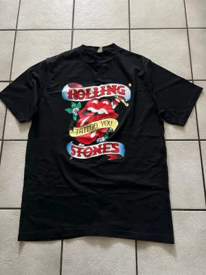 Rolling Stones svart t-shirt Tattoo You storlek XL, bomull  - Svart t-shirt med färgglatt tryck av Rolling Stones klassiska tunga och texten 'Tattoo You'. T-shirten har korta ärmar och rund hals. Perfekt för dig som gillar bandmerch och vill sticka ut med en ikonisk design.