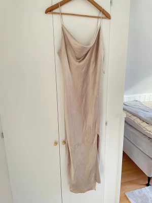 Beige långklänning med slits - Säljer en elegant beige långklänning med tunna axelband och draperad urringning. Klänningen har en slits på sidan och är tillverkad i ett glansigt material som ger en lyxig känsla. Perfekt för sommarens alla tillfällen.
