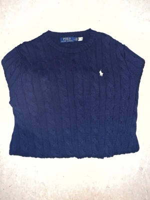 Mörkblå kabelstickad tröja Polo Ralph Lauren - Kabelstickad mörkblå tröja från Polo Ralph Lauren med rund halsringning och klassisk vit broderad logga på bröstet. Tröjan har långa ärmar och ribbade muddar vid ärmslut och nederkant. Perfekt för en stilren och tidlös look.