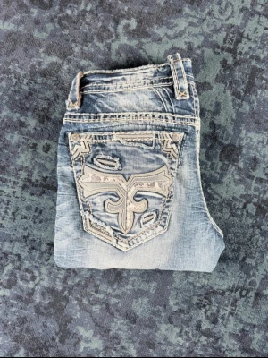 *SUPER RARE* Rock Revival Jeans - Säljer ett par ljusblå Rock Revival jeans med snygga slitningar och broderad bakficka i vitt och grått. Jeansen har raka ben, låg midja och detaljerade sömmar som ger en cool och unik look. Perfekta för dig som gillar streetstyle och vill sticka ut.