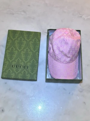 Rosa keps från Gucci med monogram - Säljer en snygg rosa keps från Gucci med klassiskt GG-monogram i hela tyget. Kepsen har böjd skärm och är tillverkad i ett glansigt tyg som ger en lyxig känsla. Perfekt accessoar för dig som vill sticka ut och addera lite designer-vibe till din outfit.