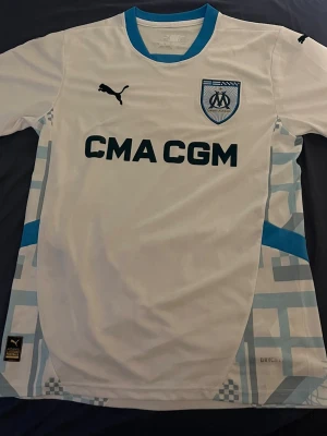 Marseille matchtröja Puma vit/blå - Säljer en vit Olympique de Marseille matchtröja från Puma med blå detaljer och klubbmärke på bröstet. Tröjan har sponsortryck 'CMA CGM' och coola grafiska mönster i ljusblått på sidorna. 