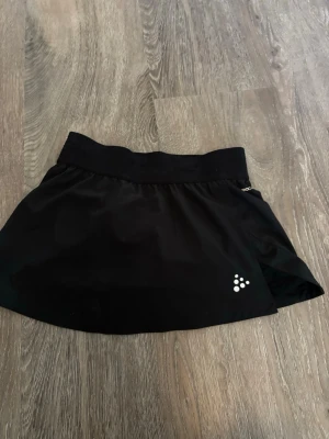 Svart sportig kjol från Craft - Säljer en svart sportig kjol från Craft med bred resår i midjan och diskret logga framtill. Kjolen har en klassisk A-linjeform och är tillverkad i ett lätt, stretchigt material som passar perfekt för träning eller aktiva dagar. Kjolen har undershorts där det finns en ficka för att stoppa bollen i