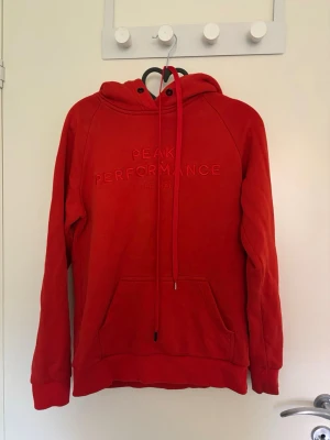 Röd/orange hoodie från Peak Performance - Snygg röd hoodie från Peak Performance med broderad logga på bröstet. Klassisk känguruficka framtill, ribbade muddar och justerbar huva med dragsko. Perfekt för en avslappnad och sportig stil. Är storlek m men sitter som en s