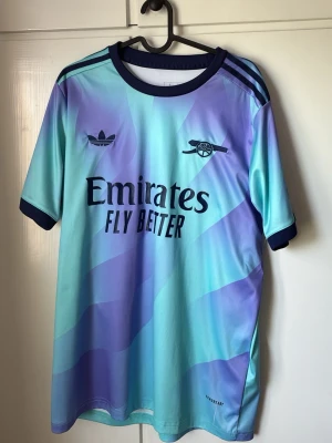 Arsenal x Adidas retro matchtröja - Snygg Arsenal matchtröja i retrostil från Adidas med blå och lila gradient, mörkblå detaljer och klubbens logga på bröstet. Tröjan har korta ärmar, rund hals och tryck som 'Emirates Fly Better' och 'Visit Rwanda'. Perfekt för fotbollsfans! lite slitage men inget som påverkar tröjan! 