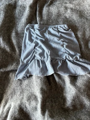 Blå kortkjol med volang - Söt blå kortkjol från Gina Tricot med tunna vita ränder och volangkant. Kjolen har rynkade detaljer framtill som ger en snygg formad look. 