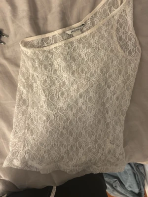 Vit spets oneshoulder topp Monki - Säljer en vit oneshoulder topp från Monki i genombruten spets. Toppen är ärmlös och har ett elegant spetsmönster som ger en romantisk vibe. Perfekt att styla med jeans eller kjol för en trendig look.