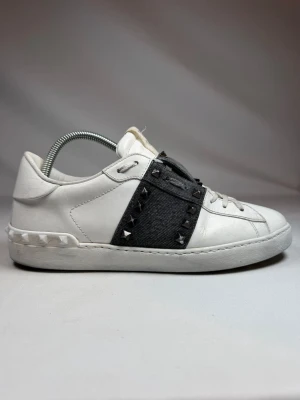 Valentino Rockstud Untitled sneakers - Snygga vita sneakers från Valentino med svart band över mitten dekorerat med silverfärgade nitar. Skorna har en låg profil och är tillverkade i läder med coola detaljer på sulan. Perfekta för dig som vill sticka ut med en edgy touch.