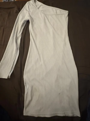 Ljusblå ribbad oneshoulder klänning - Ljusblå ribbad klänning med oneshoulder-design och lång ärm. Klänningen är figurnära och har en mjuk, stretchig känsla som gör den bekväm att bära. Perfekt för dig som vill sticka ut med en unik och stilren look. Se bild på fläck, ej testat få bort den. 
