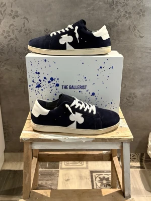 Marinblå sneakers från The Gallerist - Snygga marinblå sneakers i mocka från The Gallerist med vita detaljer och vita skosnören. Skorna har en låg profil. Skorna är storlek 44 och använda ett fåtal gånger.