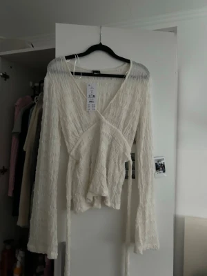 Vit omlottopp med knyt från Gina tricot  - Trendig vit omlottopp från Gina tricot med långa ärmar och knytband i midjan. Toppen har en v-ringad design och är tillverkad i ett lätt, strukturerat material som ger en cool och luftig känsla. Perfekt för att styla med jeans eller kjol.