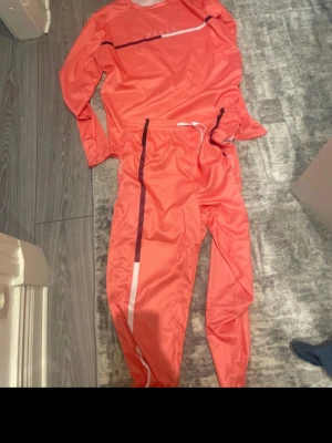 Korallfärgad träningsdress Paris - Säljer en snygg korallfärgad träningsdress från Paris. Setet består av långärmad tröja och matchande byxor med svarta och vita detaljer. Materialet är lätt och andas, perfekt för träning eller chill. Tröjan har tryckt logga och byxorna har resår i midjan.