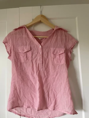 Rosa kortärmad skjorta med knappar - Säljer en rosa kortärmad skjorta med v-ringning och fyra knappar framtill. Skjortan har två bröstfickor med lock och en avslappnad passform. Perfekt till jeans eller shorts för en chill look.