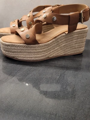Oanvända Bruna platåsandaler/Espadrillos med nitar och kilklackfrån H&M - Säljer ett par bruna sandaler med breda remmar i skinnimitation och silverfärgade nitar. Sandalerna har hög platåsula i flätad jute och justerbart spänne vid ankeln. Perfekta för dig som vill ha både höjd och stil.Klackhöjd 8 cm.