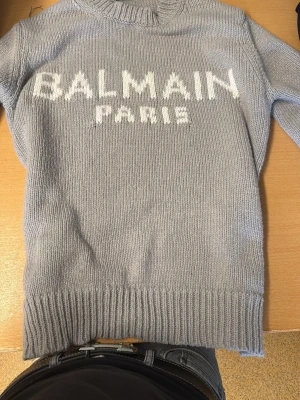 Ljusgrå stickad tröja Balmain Paris - Snygg ljusgrå stickad tröja från Balmain Paris med vit logotyp framtill. Tröjan har rund hals, långa ärmar och ribbstickade muddar vid ärmslut och nederkant. Perfekt för dig som gillar exklusiva märken och enkel, stilren design.