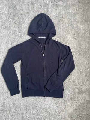 Mörkblå hoodie från RYVLS Stockholm - Mörkblå hoodie från RYVLS Stockholm med dragkedja framtill och två fickor. Tillverkad i mjukt stickat material och har ribbade muddar vid ärmslut och nederkant. Perfekt för en avslappnad och stilren look.