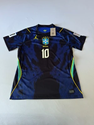 Brasilien Vini Jr. matchtröja blå - Snygg blå och svart Brasilien matchtröja från Jordan med gula detaljer och nummer 10. På framsidan finns Brasil-logga och på baksidan står det VINI JR. Tröjan har korta ärmar och är tillverkad i ett lätt, ventilerande material perfekt för fotboll.