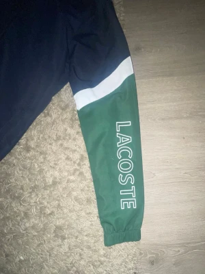 Lacoste blågrön tracksuit set - Tvådelad tracksuit från Lacoste i marinblått och grönt med vita detaljer. Jackan har huva, dragkedja och stor Lacoste-logga på ärmen. Byxorna har matchande färgblock och liten logga. Materialet är lätt och sportigt, perfekt för chill eller lounge.