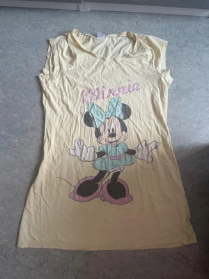 Gult nattlinne med Minnie Mouse Disney - Sött nattlinne i ljusgul färg med stort tryck av Minnie Mouse i blå klänning och rosett framtill. Mjuk och tunn bomullskvalitet, kortärmad modell med rund hals. Perfekt för dig som älskar Disney och vill ha något bekvämt att sova i.