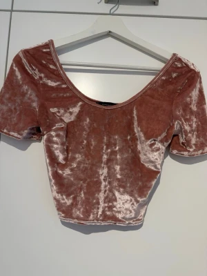 Rosa croppad sammets-topp - Snygg croppad topp i ljusrosa sammet med korta ärmar och djup urringning. Perfekt för dig som vill ha en trendig och mjuk topp med lite glansig finish. Passar till både jeans och kjol för en cool look.