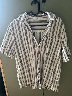 Randig skjorta från H&M i bomull - Säljer en vit skjorta med svarta vertikala ränder från H&M. Skjortan är kortärmad, har en bröstficka och knäppning framtill. Tillverkad i luftig bomull, perfekt för varma dagar. Enkel och stilren design som passar till det mesta.