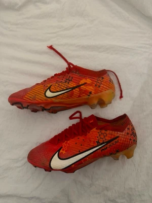 Fotbollsskor Nike ZOOM VAPOR 15 (storlek 41) - Säljer ett par röd/vit/orange Nike AIR ZOOM MERCURIAL VAPOR 15 ELITE FG DREAM SPEED 7 - fotbollsskor med orange och svart mönster samt vit swoosh på sidan. Skorna har snörning och är tillverkade i syntetmaterial med en lätt och smidig form som passar för snabba spelare. Perfekta för gräsplaner och matcher, nypris ca 3200.