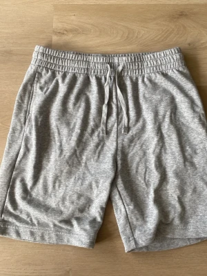 Grå mjukisshorts med snörning - Säljer ett par grå mjukisshorts med elastisk midja och snörning framtill. Shortsen har en avslappnad passform och är tillverkade i mjukt bomullsmaterial, perfekta för chill dagar eller träning.