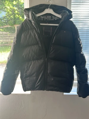 Svart pufferjacka från Tommy Hilfiger - Säljer en svart pufferjacka från Tommy Hilfiger med huva och dragkedja framtill. Jackan har diskret logga på bröstet och breda, vadderade paneler som ger en riktigt fet vinterlook. Perfekt för kalla dagar när du vill hålla värmen och ändå se stilren ut.