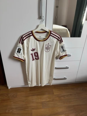 Spanien matchtröja Lamine Yamal #19 - Säljer en officiell Spanien matchtröja från Adidas med Lamine Yamal och nummer 19 på ryggen. Tröjan är vit med vinröda detaljer, tre ränder på axlarna och VM-patchar på ärmarna. Tillverkad i lätt och ventilerande material, perfekt för fotboll eller träning.