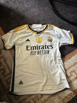 Real Madrid matchtröja Adidas - Säljer en vit Real Madrid matchtröja från Adidas med svarta och gula detaljer. Tröjan har tryck med Emirates Fly Better på framsidan och V. NI JR. samt nummer 7 på ryggen. Materialet är lätt och andas, perfekt för fotboll eller träning.