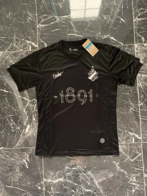 Svart AIK matchtröja Nike 1891 - Snygg svart matchtröja från Nike för AIK med trycket '1891' på bröstet och klubbens emblem. Tillverkad i ett lätt och ventilerande funktionsmaterial, perfekt för träning eller match. Kort ärm och rund hals. Modern och stilren design.
