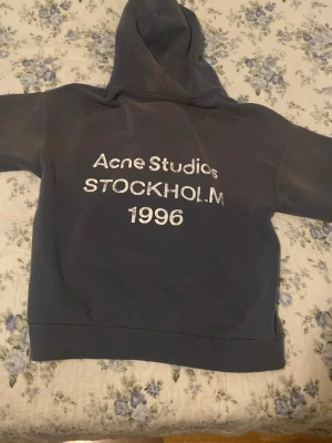 Mörkgrå hoodie Acne Studios - Mörkgrå hoodie från Acne Studios med stor huva och tryck på ryggen där det står 'Acne Studios STOCKHOLM 1996' i vitt. Oversized passform med ribbade muddar vid ärmslut och nederkant. Perfekt för en avslappnad och trendig stil.