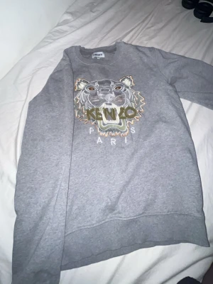Grå sweatshirt från Kenzo med tiger - Säljer en grå sweatshirt från Kenzo med det ikoniska tigerhuvudet broderat framtill och texten 'Kenzo Paris' under. Tröjan har rund halsringning, långa ärmar och är gjord i mjukt bomullsmaterial. Perfekt för dig som gillar streetwear och vill sticka ut.