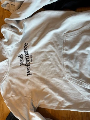 Vit hoodie från Peak Performance - Säljer en vit hoodie från Peak Performance med stor logga och text 'Est. 1986' på bröstet. Hoodien har en klassisk känguruficka framtill och är tillverkad i mjukt bomullsmaterial. Perfekt för dig som gillar stilren och sportig look.den är en Xs