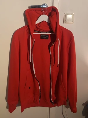 Röd hoodie från Ombre - Röd hoodie från Ombre med dragkedja framtill, vita dragsnören i huvan och två stora fickor. Klassisk modell med ribbade muddar vid ärmslut och nederkant. Perfekt för en avslappnad och cool stil.
