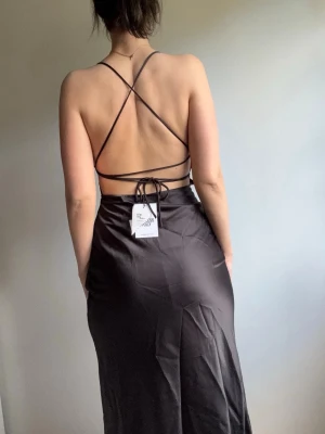 Svart långklänning med öppen rygg - Snygg svart långklänning i satinliknande material med tunna axelband och öppen rygg med snörning. Klänningen har en figurnära passform och slits baktill. Perfekt för dig som vill ha en elegant och trendig look.