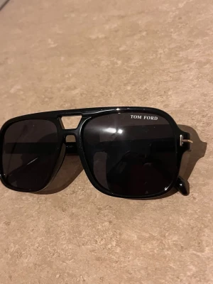 Svarta Tom Ford pilotbågar solglasögon - Snygga svarta solglasögon från Tom Ford med klassisk pilotform och mörka glas. Bågarna har en cool detalj med guldfärgad T-logga vid gångjärnen och robust plastmaterial. Perfekta för dig som vill ha en stilren och trendig look.