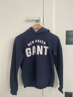Mörkblå hoodie från GANT - Snygg mörkblå hoodie från GANT med vit text 'NEW HAVEN GANT' . Klassisk huva och ribbade muddar. Perfekt för en avslappnad  stil. Mjuk bomullsblandning som känns skön mot huden.
