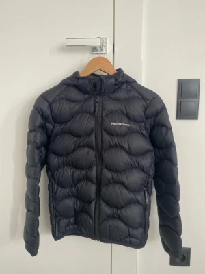 Svart dunjacka från Peak Performance - Snygg svart dunjacka från Peak Performance med huva och dragkedja framtill. Jackan har två sidofickor med dragkedja och diskret logga på bröstet. Puffig design med vågigt mönster och lätt material som håller dig varm under kalla dagar.