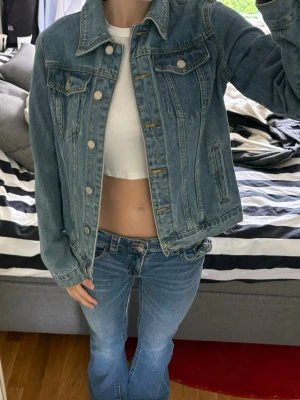 Klassisk blå jeansjacka - Säljer en klassisk blå jeansjacka med raka sömmar och silverfärgade knappar. Jackan har två bröstfickor med lock och knapp samt två sidofickor. Tidlös modell med krage och justerbar nederkant. Perfekt att slänga över en t-shirt för en avslappnad look.