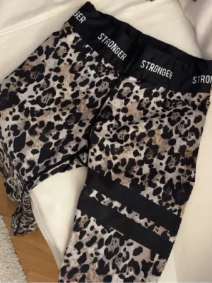 Leopardmönstrade leggings från Stronger - Snygga leopardmönstrade leggings från Stronger med bred resår i midjan där loggan syns tydligt. Leggingsen har svarta detaljer runt låren och är tillverkade i ett stretchigt funktionsmaterial som passar perfekt till träning.