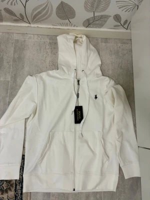 Vit hoodie från Polo Ralph Lauren - Snygg vit hoodie med dragkedja från Polo Ralph Lauren. Klassisk design med svart broderad logga på bröstet, justerbar huva med snören och två stora fickor framtill. Tillverkad i mjuk bomull för skön komfort och stilren look.